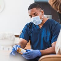 During COVID-19, a mid adult male healthcare provider visits a senior woman in her home to help her with her medications.  He is sitting at a safe distance and wears protective gloves and mask.