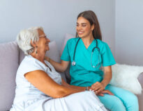 Happy patient is holding caregiver for a hand while spending time together. Elderly woman in nursing home and nurse. Aged elegant woman and tea time at nursing home