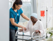 Young female home healthcare professional helps an elderly woman get out of bed by using a walker. The woman is wearing a cap on her head. An iv drip is in the background.